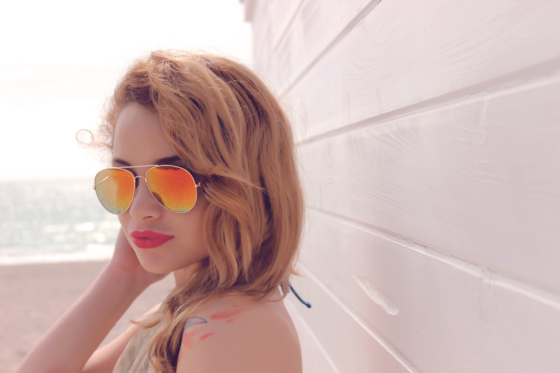 Eine junge blonde Frau mit schillernder Sonnenbrille lehnt an einer Holzwand im Sonnenschein. Welche Haarpflege sie im Sommer braucht?