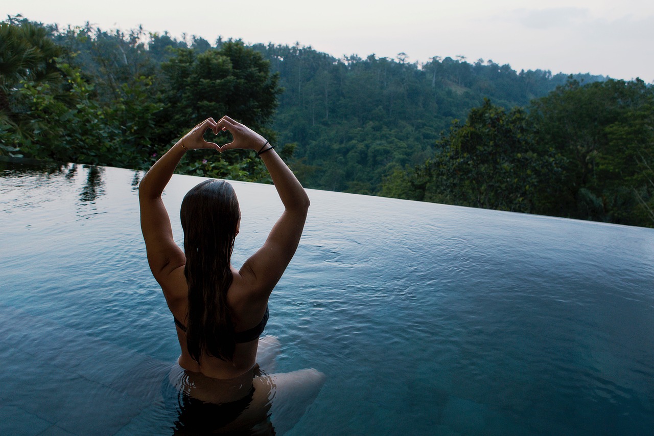 Eine Frau sitzt in einem Pool, hat die Arme über dem Kopf erhoben und schaut in ein Tal voller Bäume, während sie mit den Händen ein Herz formt.