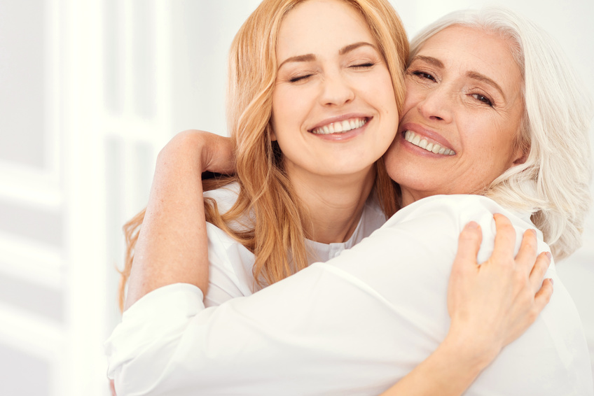 Eine junge blonde Frau umarmt eine ältere Frau mit hellgrauen Haaren. Beide sehen gepflegt aus, tragen weiße Kleidung und lächeln.