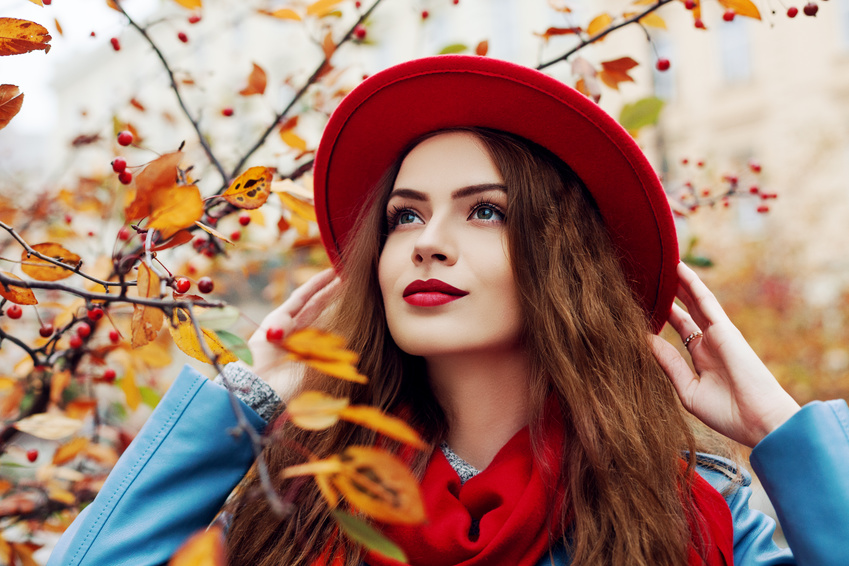 Eine Frau mit langen braunen Haaren steht vor einem Busch mit Herbstlaub. Sie trägt eine blaue Jacke, einen roten Schal und Hut, den sie mit beiden Händen festhält. Ihre Lippen sind rot, was einer der Make-Up Trends 2019 ist.