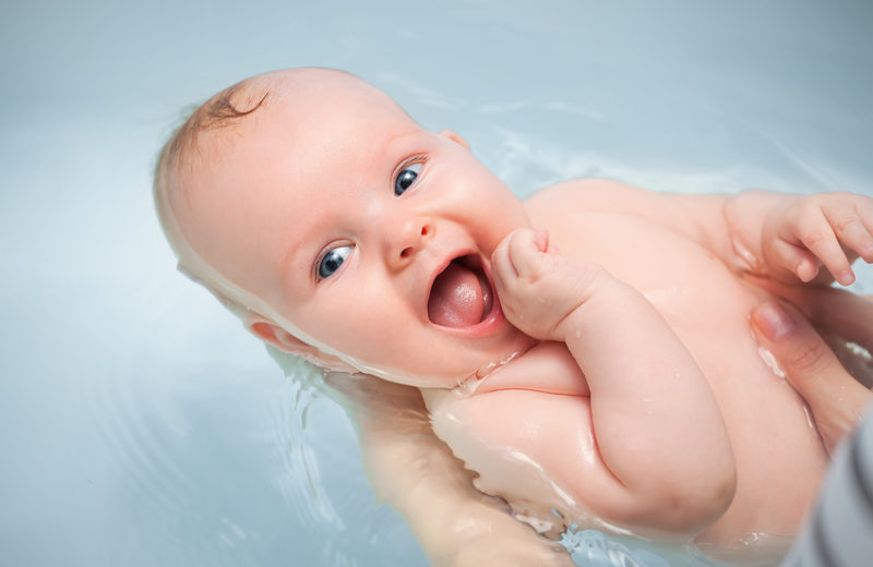 Man sieht, wie groÃe HÃ¤nde ein kleines, lachendes Baby baden. Das Wasser ist klar .